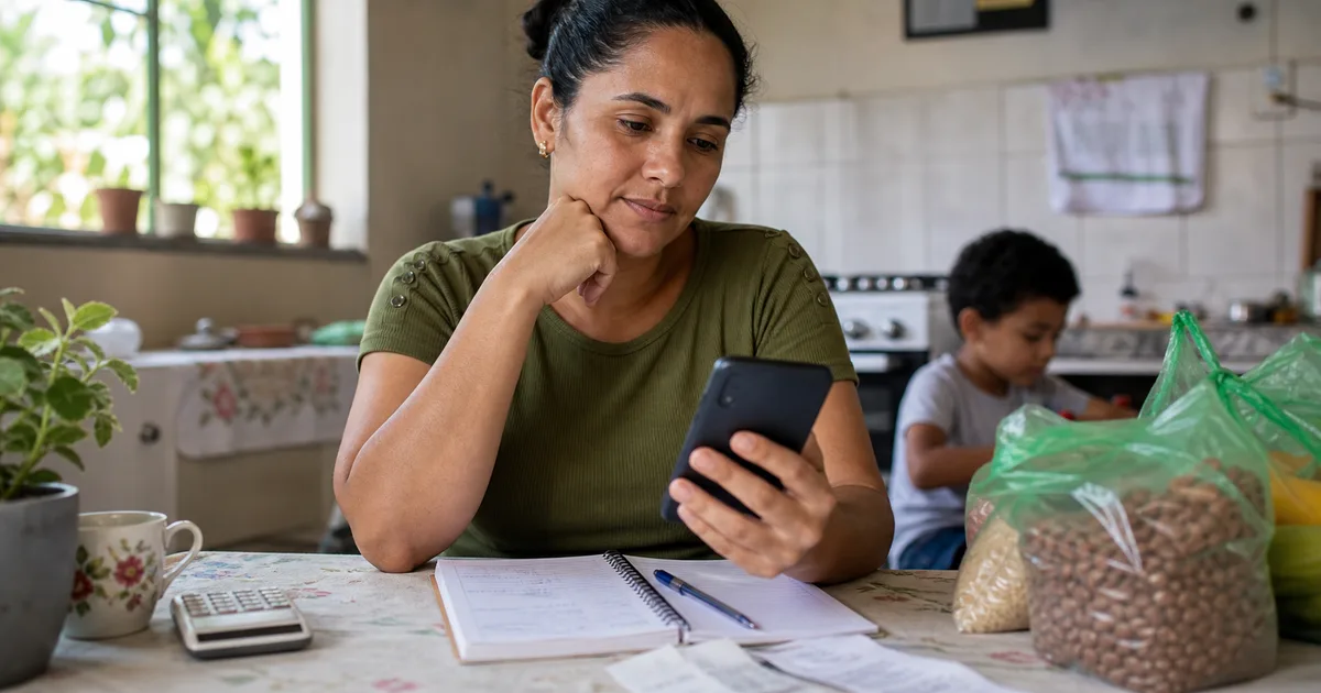 Mulher brasileira consulta no celular informações sobre o valor do Bolsa Família em 2026 ao lado de contas e compras de mercado.